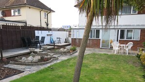 Ein Garten mit einer Steinterrasse, einem kleinen Gartenbeet und einer Sitzgruppe mit Stühlen und einem Tisch.