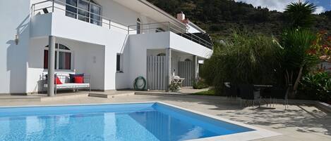 A modern two-story house with a swimming pool, outdoor furniture, and a view of a mountainous landscape.