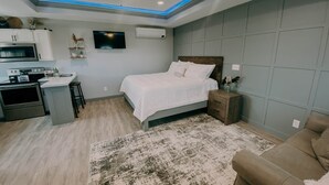 A bedroom with a bed, a nightstand, a TV, a ceiling fan, and a kitchenette. - The Honeymoon Hideaway (Trenton)