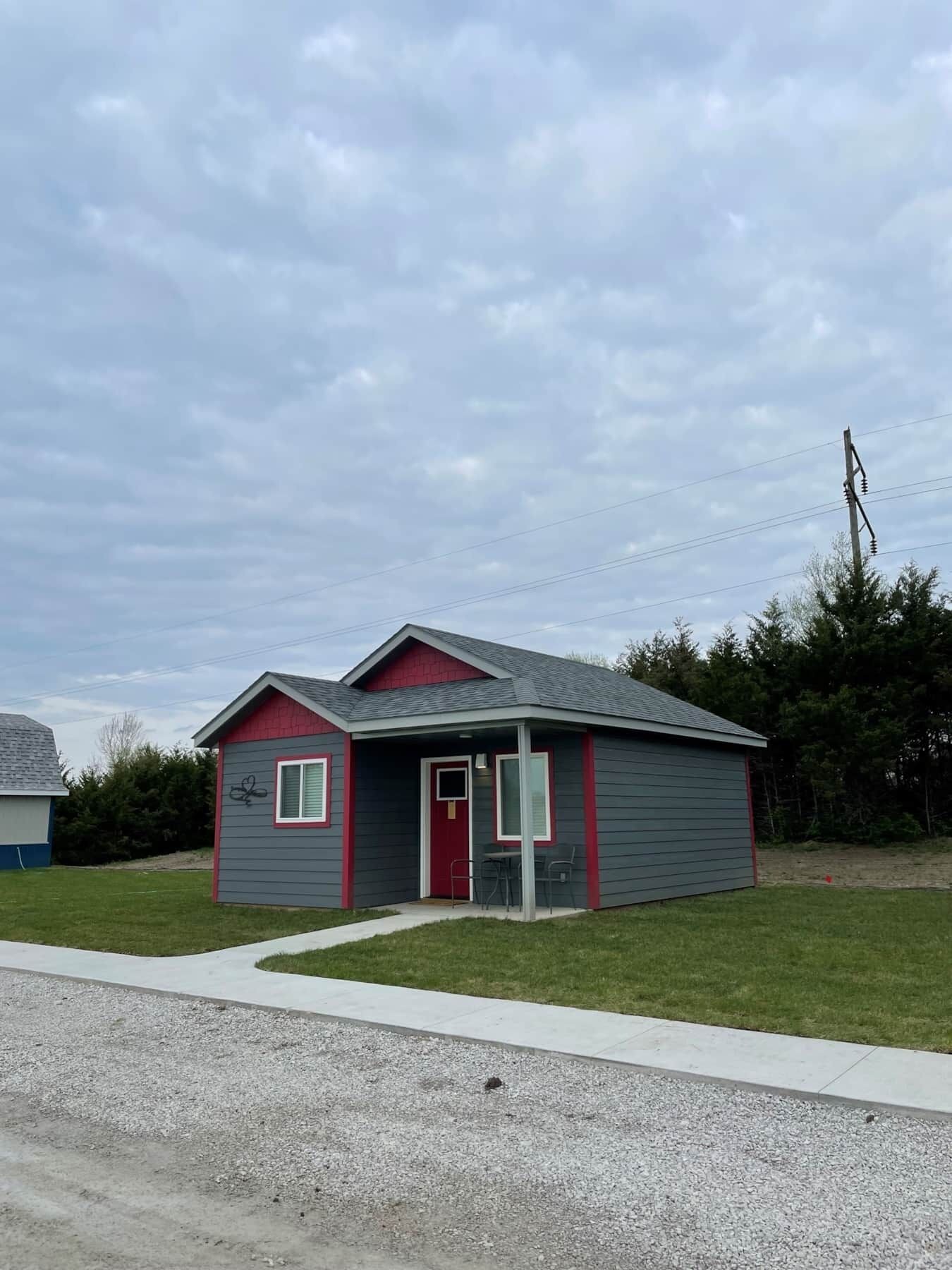A small, single-story house with a red door and grey siding, located on a grassy area with a gravel driveway.