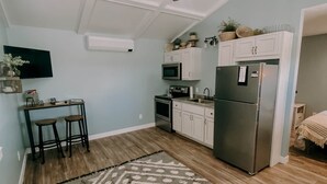 A compact kitchen with white cabinets, a stainless steel refrigerator, a microwave, and a stove. There is a patterned rug on the floor, and a hanging light fixture above the kitchen counter. - The Modern Farm House (Trenton)