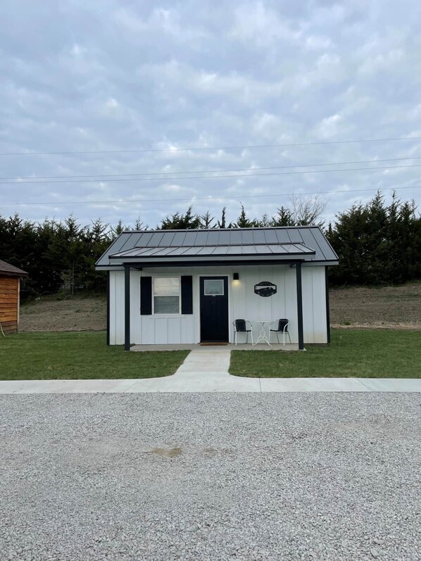 A small, single-story building with a dark roof and white exterior walls. It features a front door, two windows, and a small porch area with two chairs. - The Modern Farm House (Trenton)