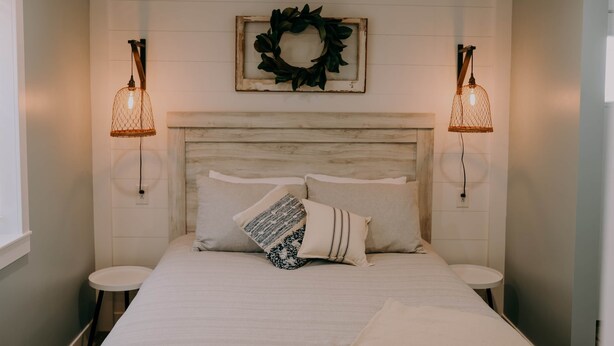 A bedroom with a wooden headboard, two bedside lamps, a decorative wreath, and a bed with patterned pillows. - The Modern Farm House (Trenton)