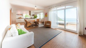 A spacious living area with a sofa, a dining table, and chairs. There is a kitchen in the background, and a sliding glass door leading to a balcony with a view of the sea.