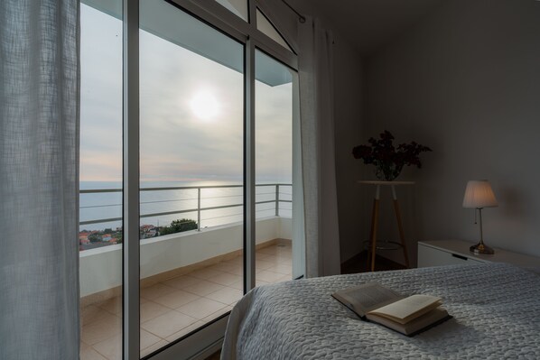 A bedroom with a large window, a bed with a book, a bedside table with a lamp, and a view of the city.