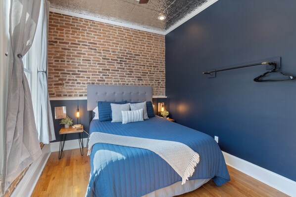 Un dormitorio con pared de ladrillo, cama azul y mesita de noche de madera con lámpara.