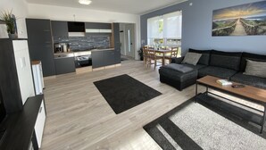 A modern kitchen with grey cabinets, a black countertop, and a built-in oven. There is a dining area with a table and chairs, and a living room with a sofa and a coffee table.