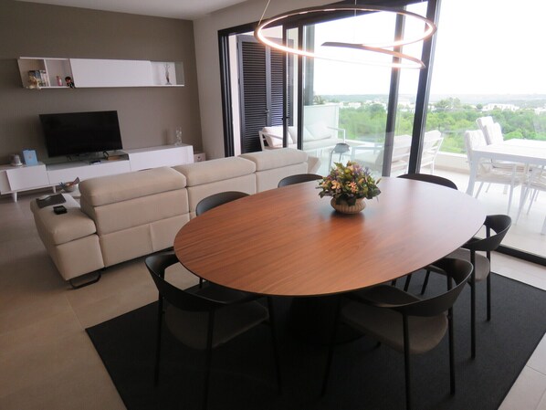 A modern living room with a round wooden dining table, beige sofa, and a TV on a white cabinet.