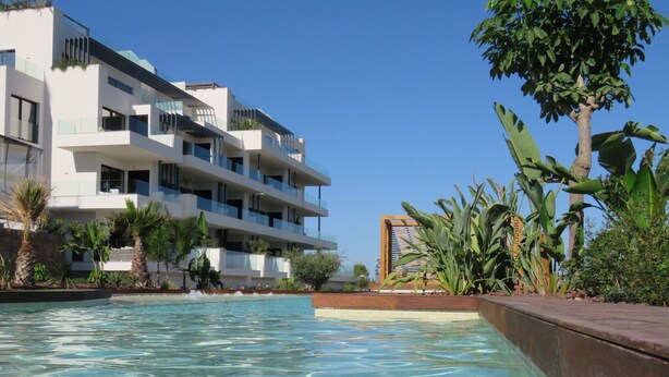 A modern hotel with a pool, surrounded by tropical plants and a clear blue sky. - Luxury south facing 3 bedroom apartment in Las Colinas Golf (Dehesa de Campoamor)