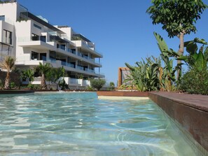 A modern hotel with a pool, surrounded by tropical plants and a clear blue sky.