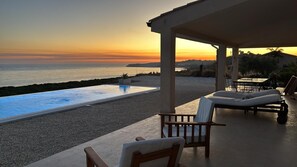 A poolside area with lounge chairs, a pergola, and a view of the sea at sunset.