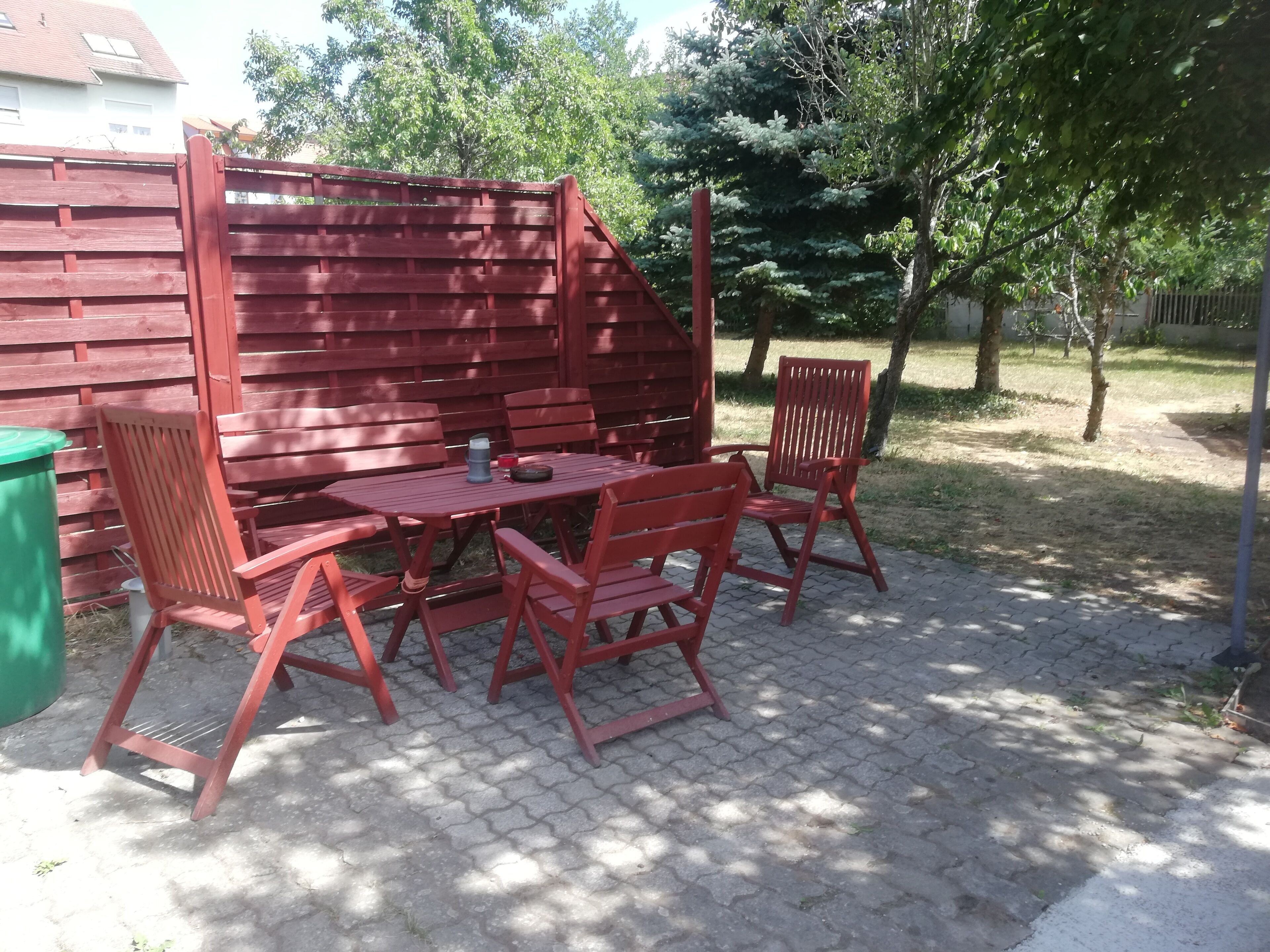 A patio with a red wooden table and chairs, a green trash bin, and a red fence.