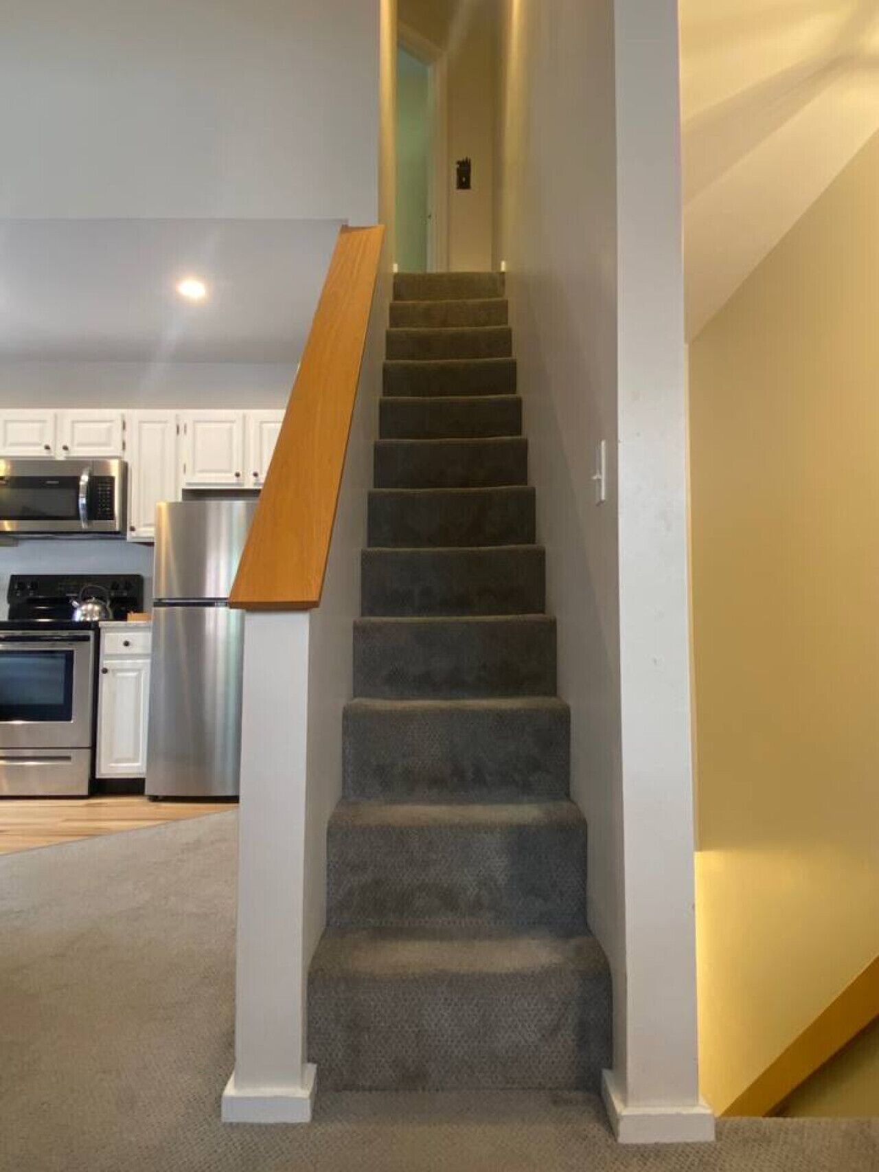 A staircase with carpeted steps and a wooden handrail, leading to a kitchen area with white cabinets and stainless steel appliances.
