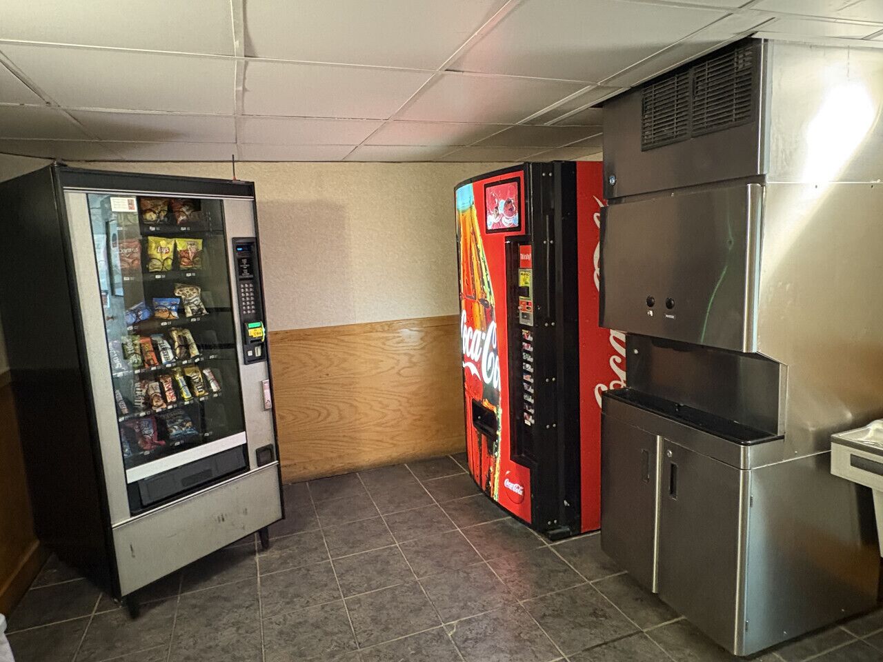 Un área con máquinas expendedoras, incluyendo una máquina de Coca-Cola, una máquina de aperitivos y un refrigerador de acero inoxidable.