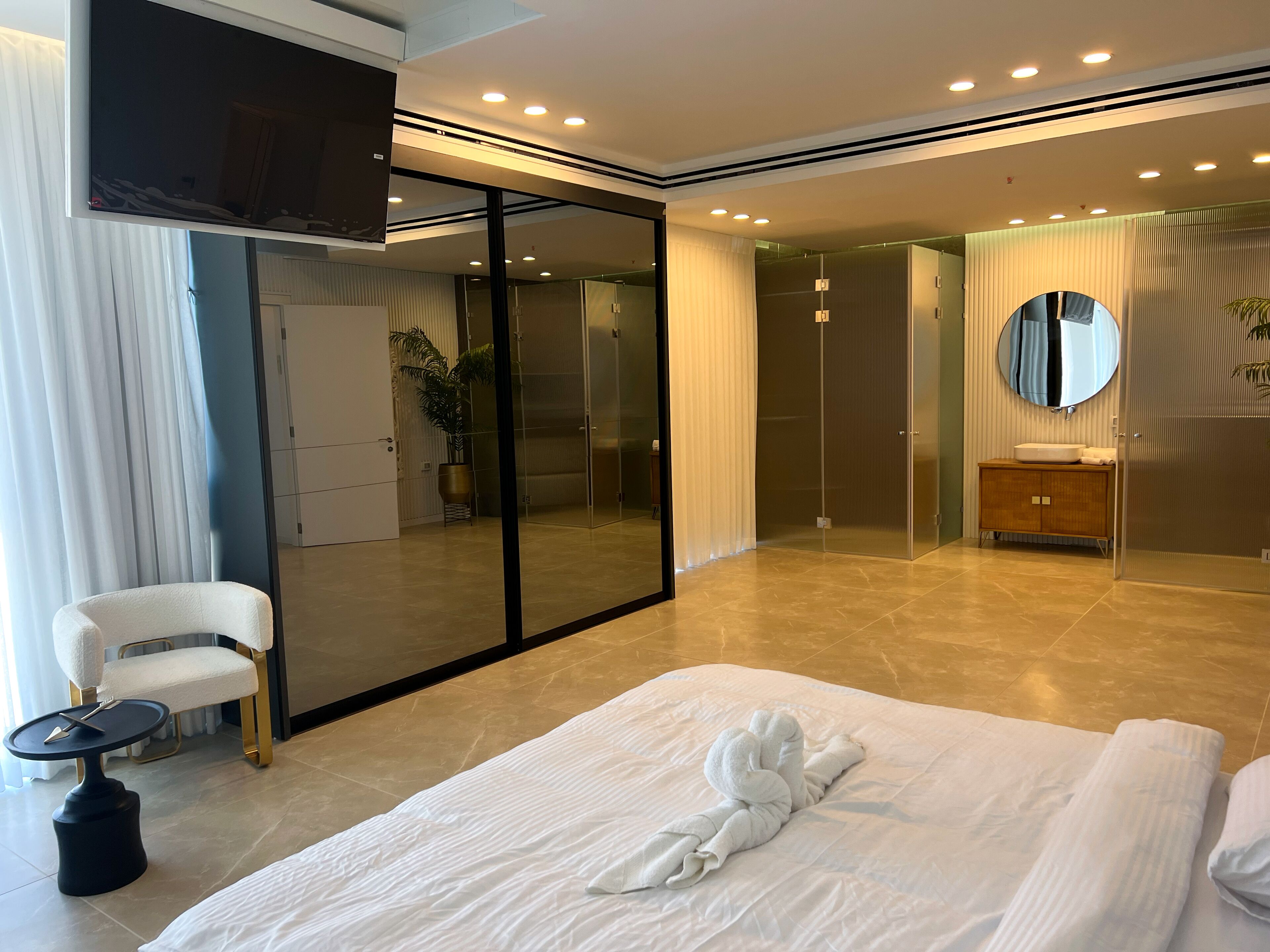 Quarto de hotel moderno com uma cama grande, área de estar com mesa, espelho e closet espaçoso.