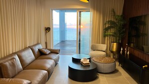 Una sala moderna con un sofá de cuero, una mesa redonda y un sillón. Hay un ventanal con vista al mar y al atardecer.