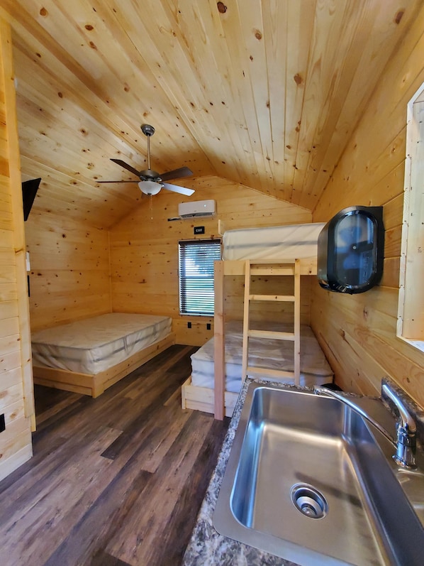 A small wooden cabin with a single bed, a built-in bench, a ceiling fan, an air conditioning unit, a sink, and a window with blinds.