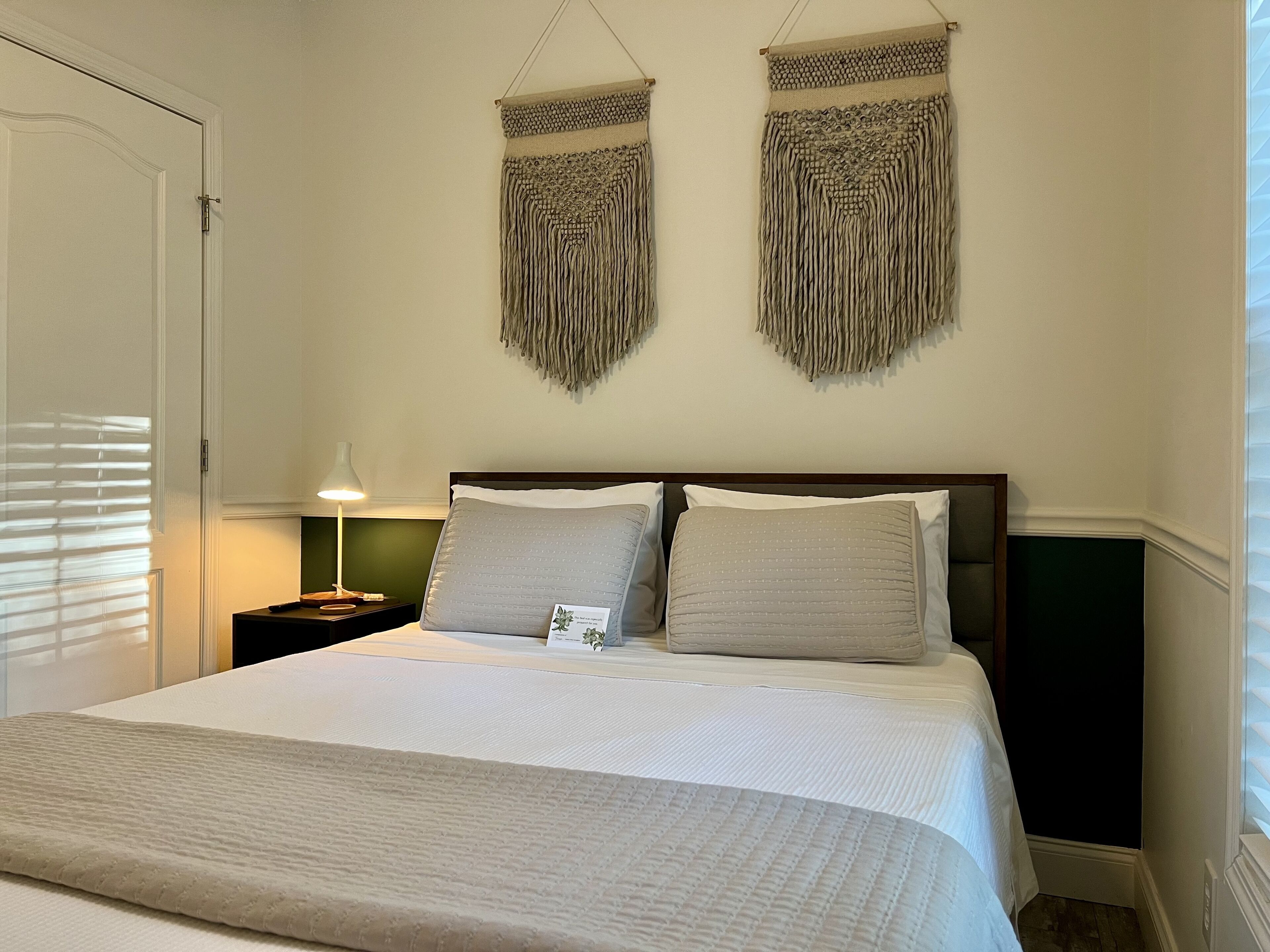 Un lit bien fait, avec une couette blanche et deux oreillers, une lampe de chevet et une décoration en macramé accrochée au mur au-dessus du lit.