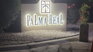 Een bord voor Palma Real met een palmboomlogo, voor een gebouw met een grindpad en een bloempot.