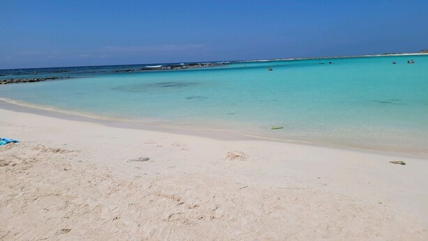 Een strand met helder turquoise water, een zandstrand en enkele mensen in de verte.