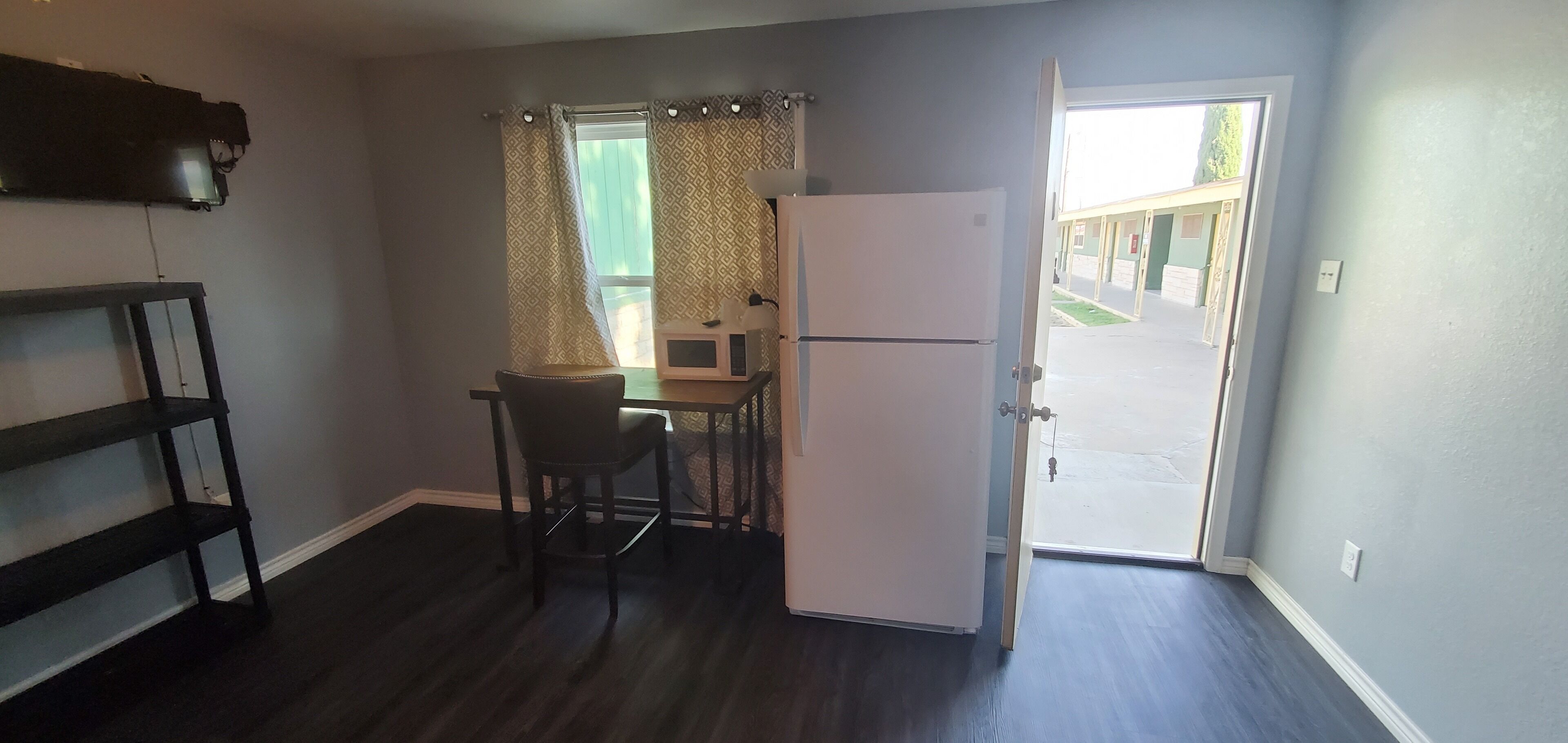 A compact kitchen with a refrigerator, a dining table, and a chair. A window with patterned curtains is visible.