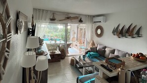 A spacious living room with a sofa, coffee table, and decorative items. There is a large window with curtains, a ceiling fan, and a wall-mounted air conditioning unit. - Quiet garden-level apartment with sea view (Sainte-Maxime)