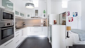 Una cucina moderna con mobili bianchi, forno integrato e frigorifero. Sul bancone ci sono oggetti decorativi e nella stanza adiacente si vede un letto con copriletto a motivi grigi e bianchi.