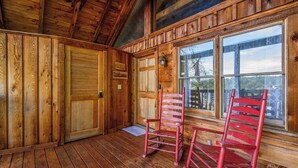 Een houten cabin interieur met een houten deur waarop 'Welcome' staat, een schommelstoel en een raam met uitzicht op bomen.