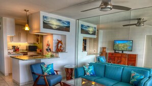 A modern living room with a teal sofa, a glass coffee table, and a kitchen area with a microwave and sink.