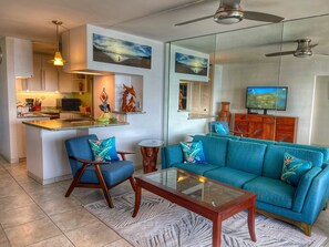 Een moderne woonkamer met een turquoise bank, een glazen salontafel en een keukenhoek met een magnetron en spoelbak.