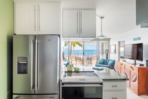 A modern kitchen with a stainless steel refrigerator, a microwave, and a coffee maker. There is a dining area with a table and chairs, and a view of the ocean through large windows. - Mermaid Cove Breathtaking Ocean Views 2 bdrm 2 bath (Wailuku)