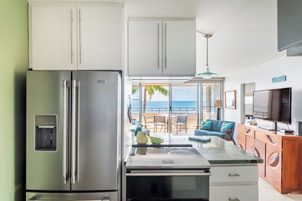 A modern kitchen with a stainless steel refrigerator, a microwave, and a coffee maker. There is a dining area with a table and chairs, and a view of the ocean through large windows.