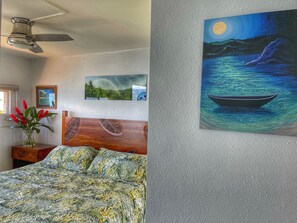 A bedroom with a bed, a ceiling fan, a night sky painting, and a framed landscape picture.