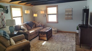A cozy living room with a leather sofa, a wooden chest, and a patterned rug. There are two wall-mounted lamps, a window with blinds, and a wall hanging.