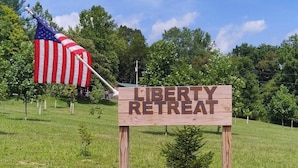 A wooden sign with "LIBERTY RETREAT" written on it, an American flag, and a grassy field with trees in the background.