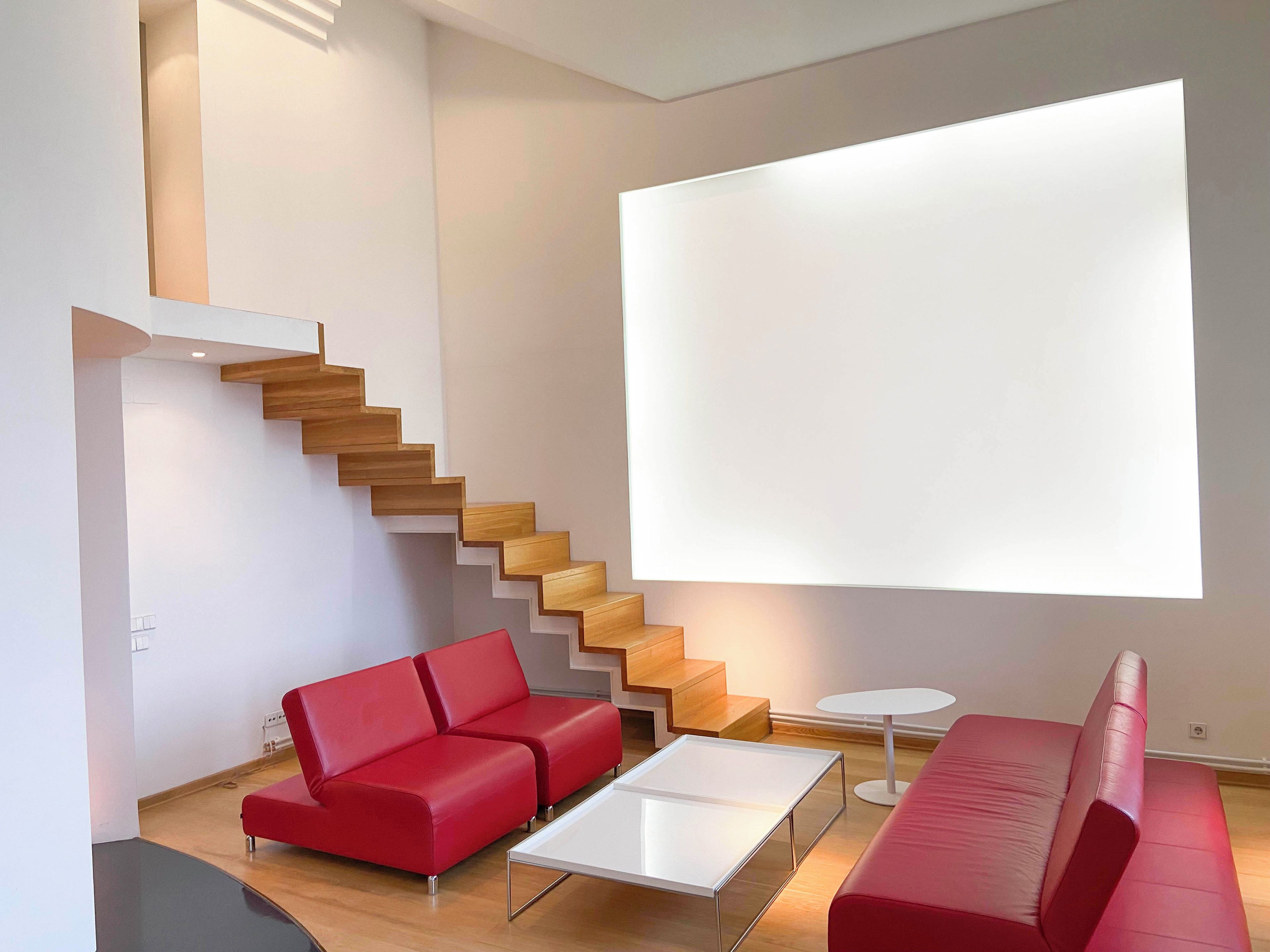 A modern living room with a staircase, red sofas, and a large window.