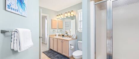 A bathroom with a light blue wall, a white toilet, a sink with a mirror above it, and a shower area.