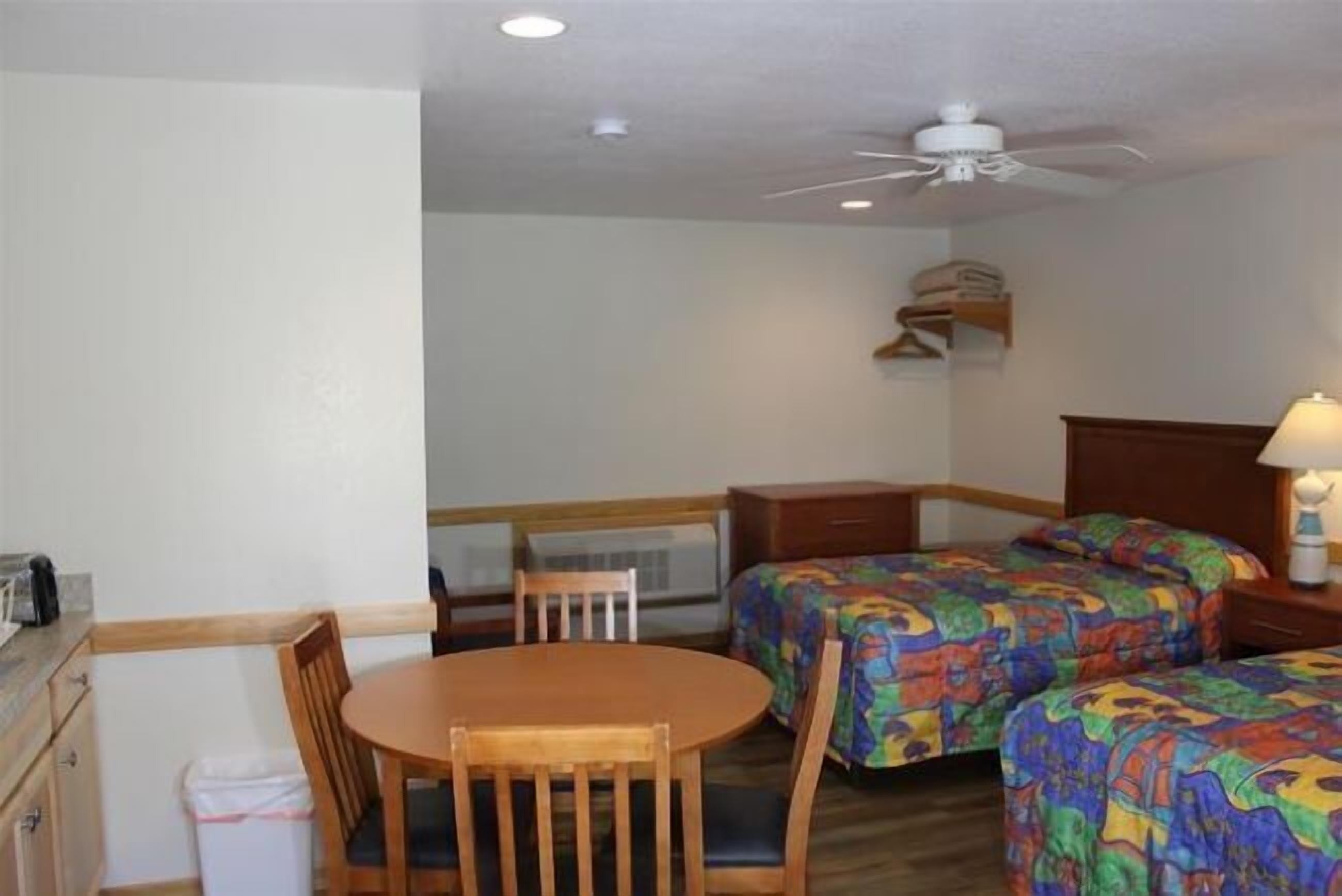 A compact hotel room with a single bed, a small table, and a chair. There is a ceiling fan and a wall-mounted shelf with a lamp.