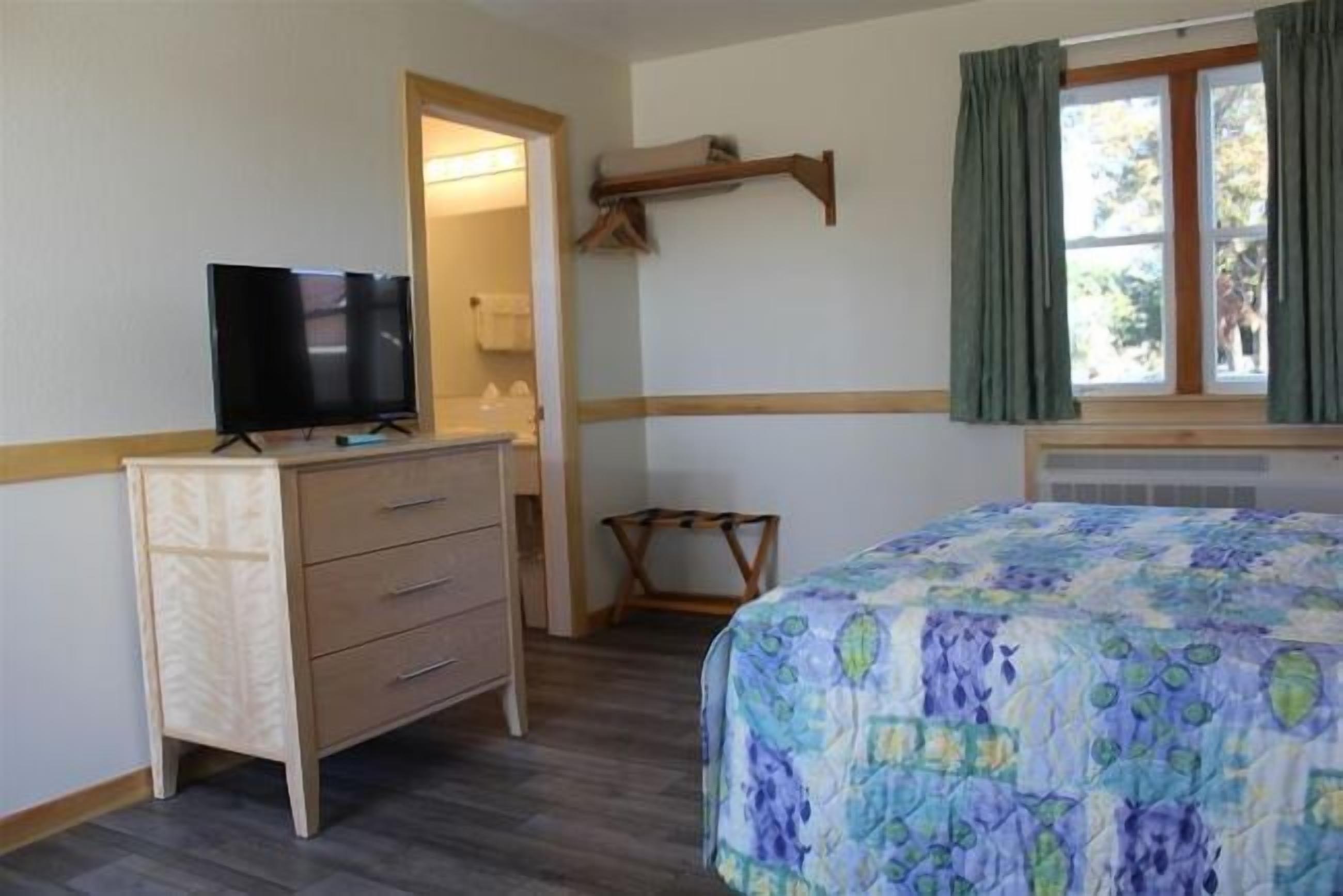 Une chambre à coucher avec un lit, une commode, un téléviseur et une fenêtre avec des rideaux.