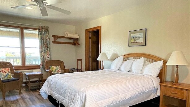 A bedroom with a large bed, two bedside tables with lamps, a chair, a small table, a ceiling fan, and a window with curtains. - Pony Island Inn (Ocracoke)