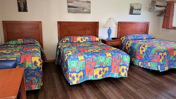 A hotel room with three wooden beds, each with colorful patterned bedding. There are wooden nightstands with lamps, and the room has wooden flooring.