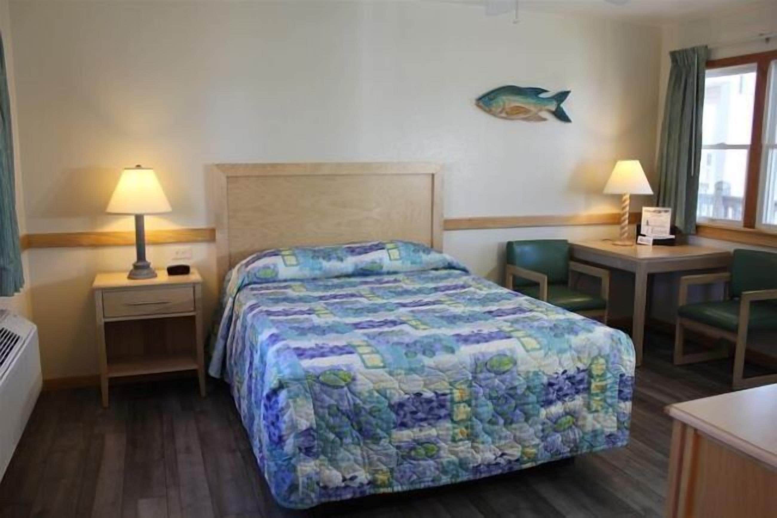 A hotel room with a bed, a desk, two chairs, a lamp, and a fish decoration on the wall.