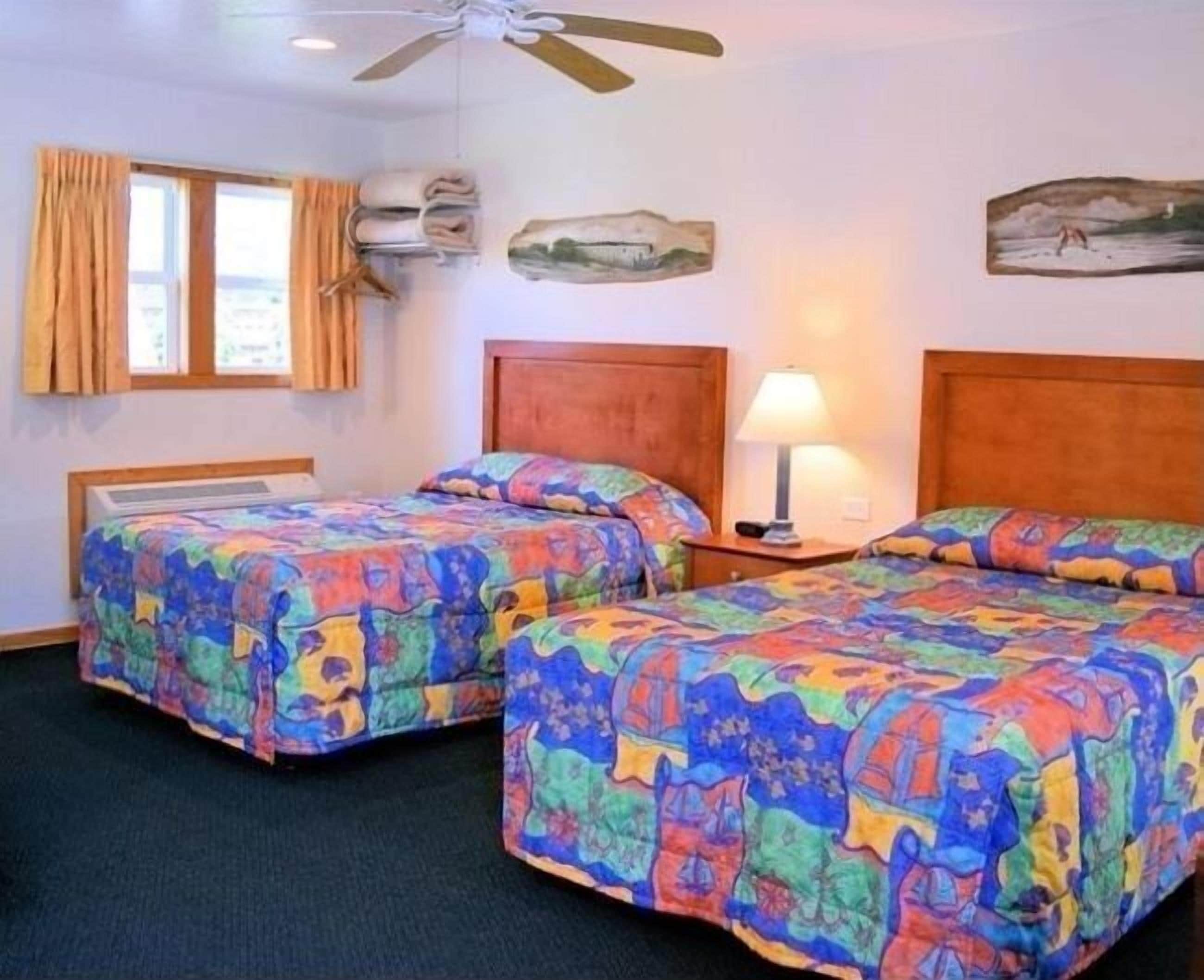A room with two beds, each with colorful patterned bedding, a ceiling fan, a window with curtains, and a wall-mounted lamp.
