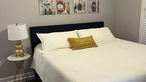 Una cama bien hecha con ropa de cama blanca y una única almohada dorada, una pequeña mesa redonda con una lámpara, y una serie de cuadros con motivos náuticos sobre la cama.