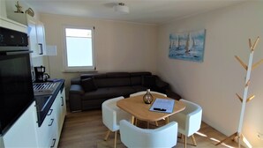 Ett kompakt bostadsutrymme med ett litet kök, en soffa, ett matbord med stolar, en tavla och en klädhängare.