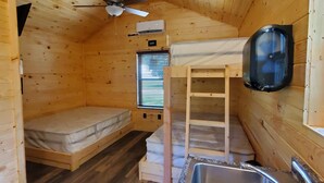 Una pequeña cabaña de madera con una cama individual, un banco integrado, un ventilador de techo, un equipo de aire acondicionado, un lavamanos y una ventana con persianas.