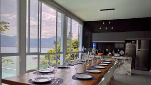 Une cuisine moderne dotée d’une longue table à manger pouvant accueillir six personnes, de grandes fenêtres offrant une vue sur la mer et les montagnes, et d’un réfrigérateur élégant en acier inoxydable.