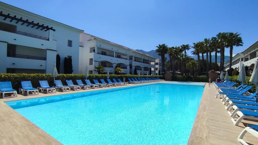 A swimming pool with lounge chairs and umbrellas, surrounded by a building with multiple balconies and palm trees.