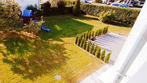 Een achtertuin met gras, een blauwe glijbaan, een barbecue en een rij jonge bomen.