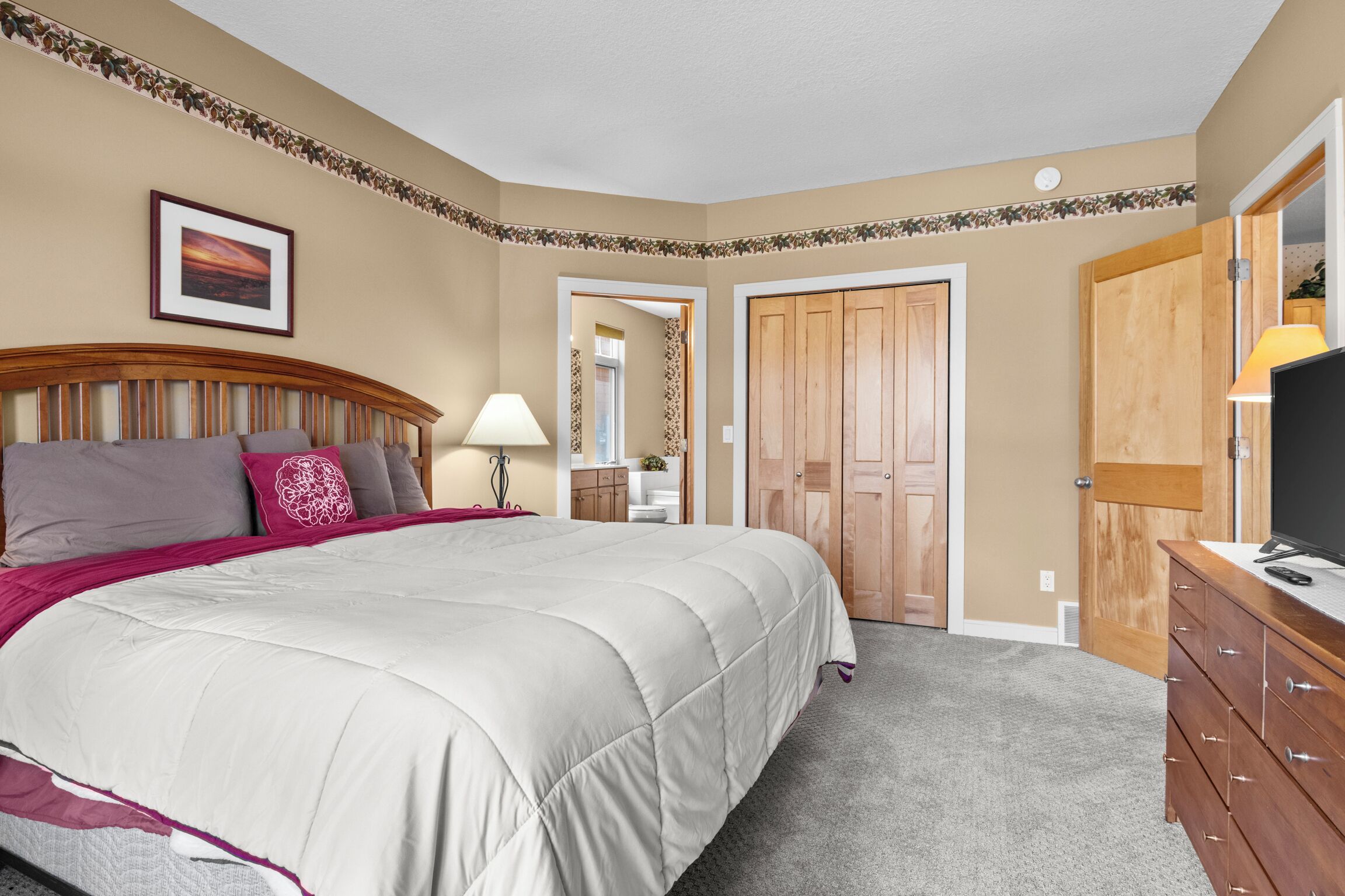 A bedroom with a bed, a dresser, a TV, and a door leading to another room.